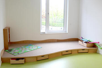 Spielecke mit grauem Teppich auf einer kleinen Erhöhung, darunter befinden sich Schubladen mit Spielsachen.