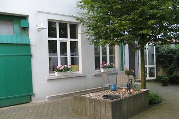 Innenhof mit Baum, Blumen, Tisch und Stühlen, im Hintergrund die Wand des Gebäudes mit Tür und Fenster