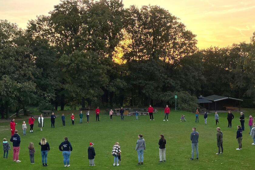 Auf einer Wiese vor einem Sonnenuntergang stehen viele Johanniter-Jugendliche in einem großen Kreis.