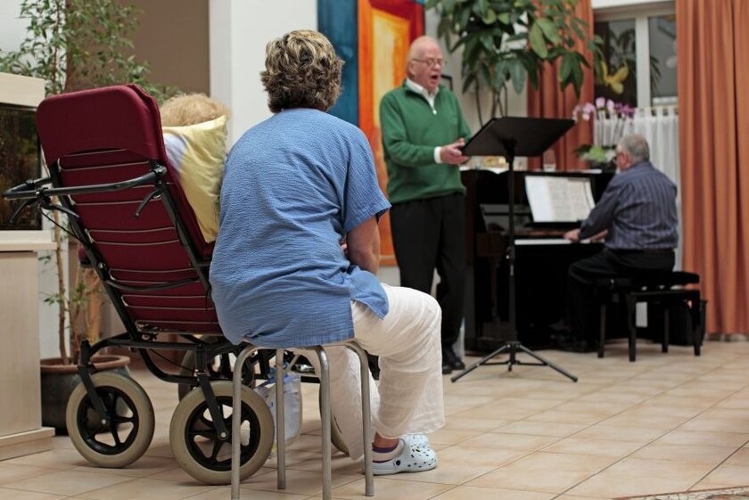 Eine Pflegerin sitzt neben einer Frau im Rollstuhl und hört einem Sänger zu.