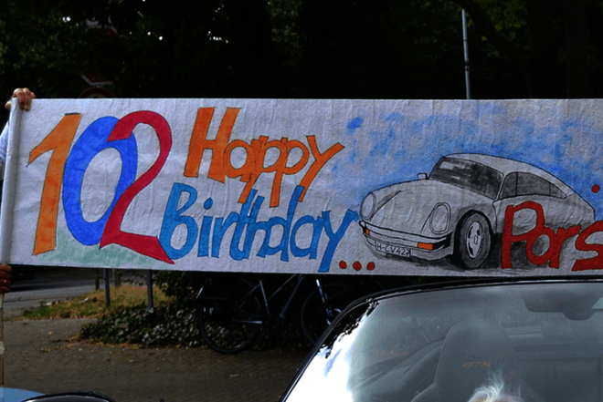 Eine Frau und ein Mann halten über das Dach eines Porsches ein Banner mit der Aufschrift "102 Happy Birthday unserer Porsche-Lady"