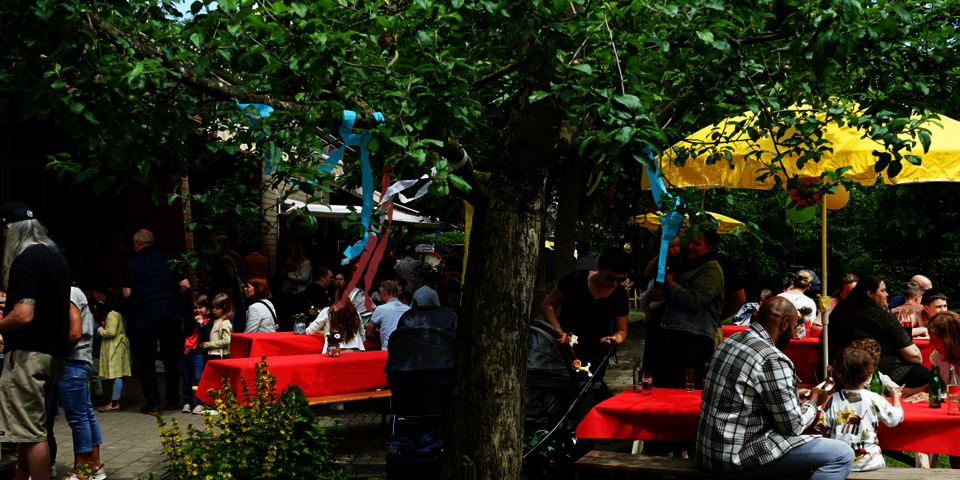 Kinder und Erwachsene sitzen im einem Garten an bunt geschmückten Tischen. Von den Bäumen hängen bunte Papierstreifen.