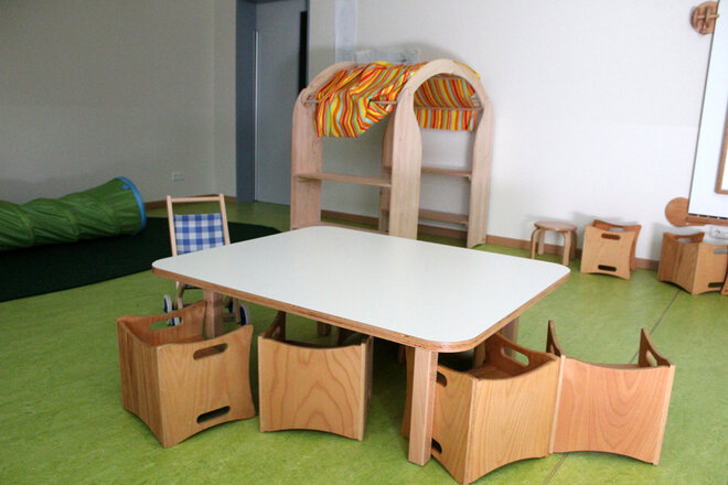 Kleiner Holztisch mit Stühlen zum Spielen. Im Hintergrund ist ein Teil der Puppenecke zu sehen. 