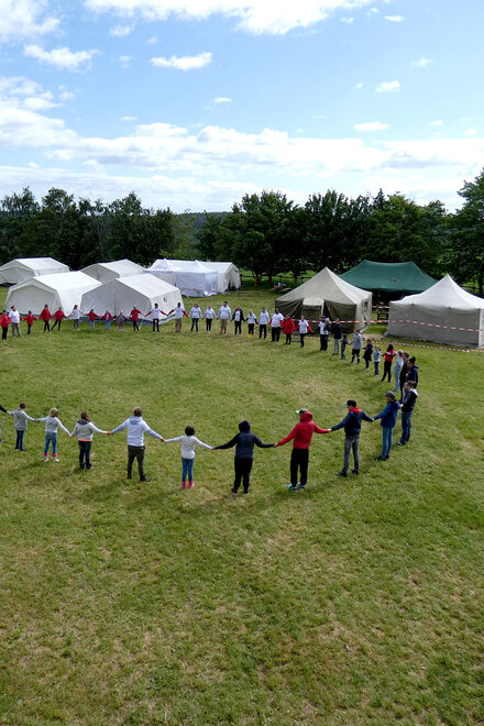 Junge Menschen stehen in einem Kreis und fassen sich an den Händen. Im Hintergrund stehen Zelte.