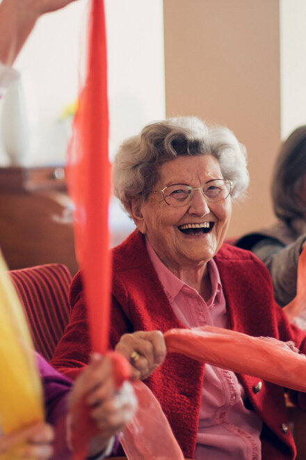 Lachende auf dem Stuhl sitzende Seniorinnen bewegen sich spielend mit einem bunten Tuch
