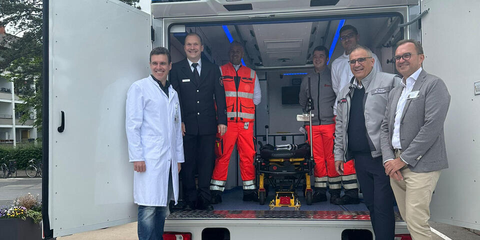 Mehrere Vertreter der Organisationen Johanniter, Feuerwehr Frankfurt, varisano Klinikum Frankfurt Höchst stehen auf der Laderampe eines Intensivtransportwagens ITW und lächeln in die Kamera.