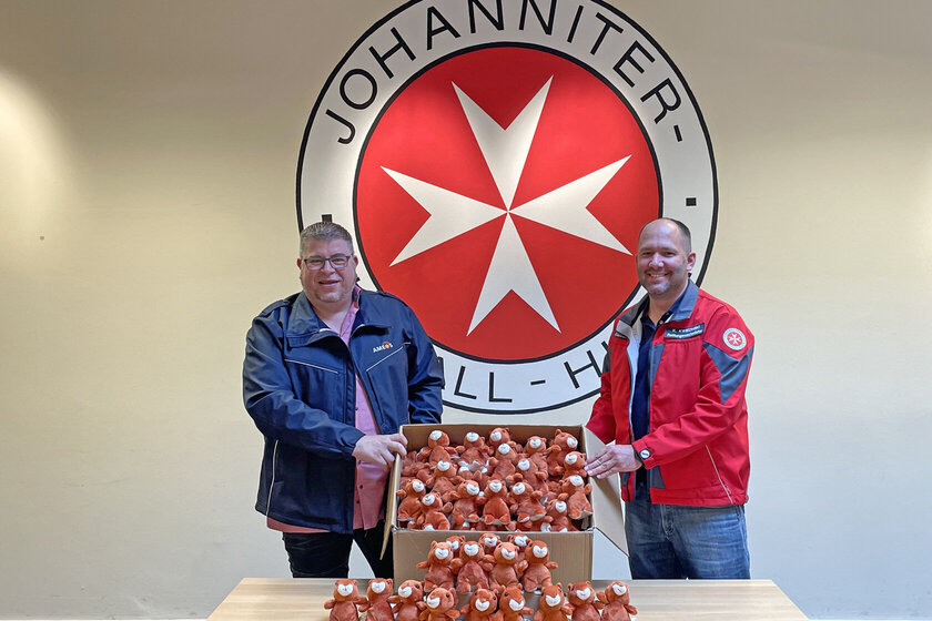2 Männer mit einem Karton voller Stoffeichhörnchen vor dem Johanniter-Logo