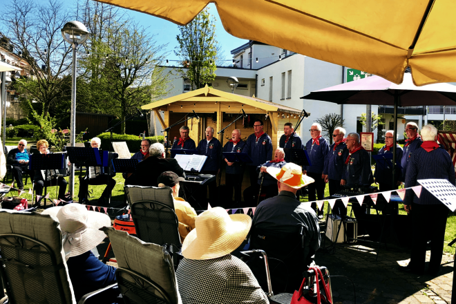 Damen und Herren mit Sonnenhüten unter einem Sonnenschirm vor eine Reihe singender Männer in blauen Seemannshemden und mit roten Halstüchern