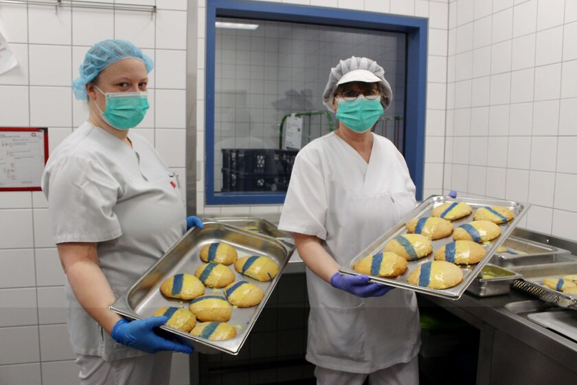 Zwei Damen mit Masken in der Küche mit einem Tablett voller Kekse in gelb-blau 