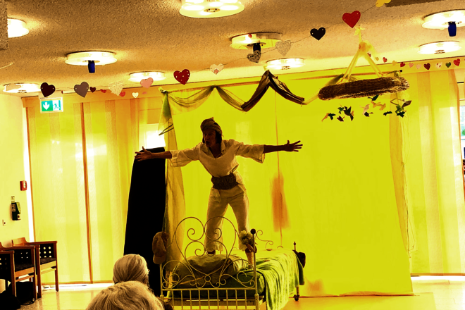 Person steht vor einem leuchtendgelben Vorhang auf einem Bett; im Vordergrund die Hinterköpfe von Zuschauenden