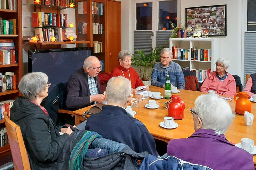 Um einen Tisch sitzen mehrere ältere Menschen, die zwei Personen zuhören, die gerade etwas vorlesen.