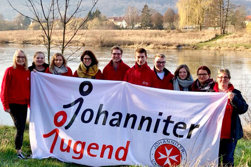 Jugendgruppenleitende der Johanniter-Jugend stehen an einem Fluss und halten ein großes Plakat mit der Aufschritt "Johanniter-Jugend". 