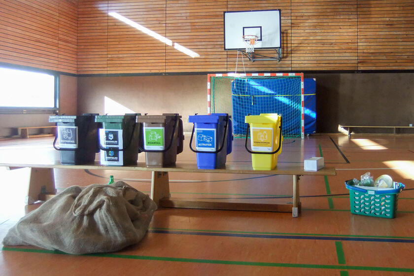 In einer Turnhalle stehen fünf Mülltonnen, ein Korb und ein Sack in verschiedenen Farben. So sieht der ideale Weg der Mülltrennung aus.