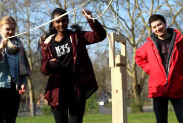 Zwie Jugendliche halten ein Seil mit dem Sie einen Turm aus Holzklötzen errichten. Ein junger Erwachsener in Johanniter-Jugend-Jacke steht im Hintergrund.