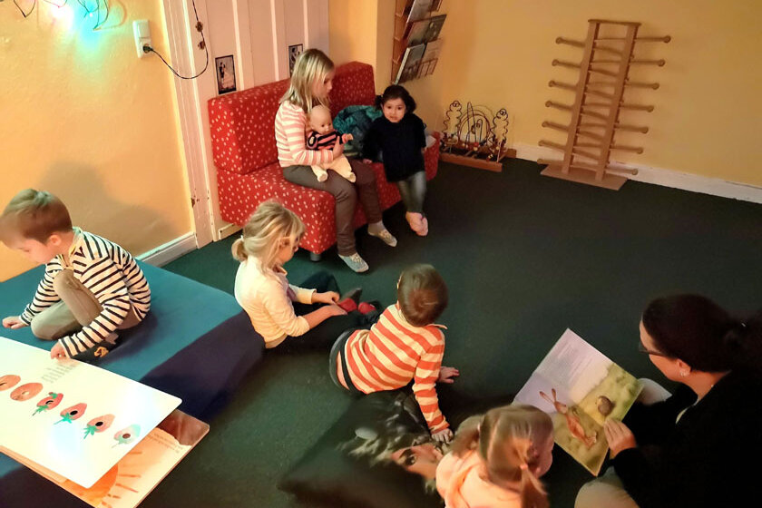 Die Kinder sitzen gemütlich im Gruppenraum und schauen sich Bücher an. An einer der Ecken sitzt eine Erzieherin, die gerade einem kleinen Mädchen vorliest.