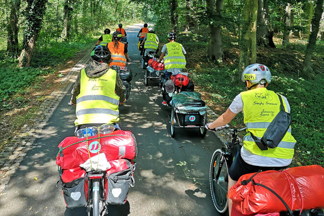 Mehrere Jugendliche auf Fahrrädern mit Helm, Warnweste und Gepäcktaschen. Sie fahren durch ein Waldstück.