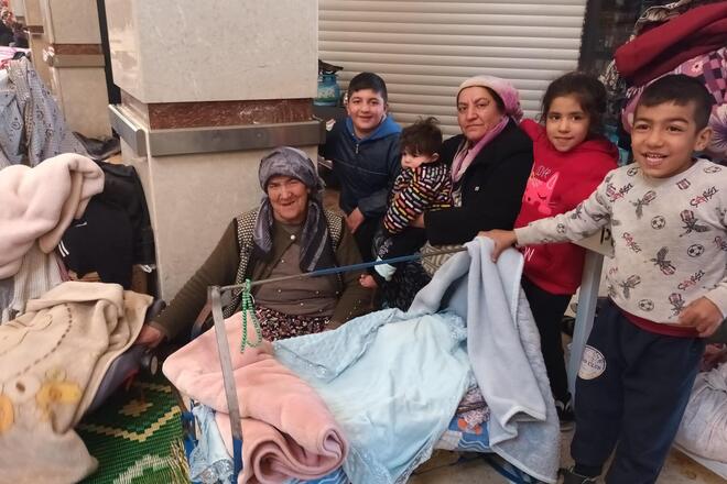 Zwei Frauen mit Kopftuch sitzen mit mehreren Kindern in einer Notunterkunft.
