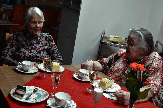 Zwei ältere Damen in gemusterten Blusen sitzen an einem gedeckten Kaffeetisch. Vor ihnen stehen Telle rmit Torte und Sektgläser. Auf dem Tisch liegt eine rote Decke; und in einer Vase steht eine orange Gerberablüte..