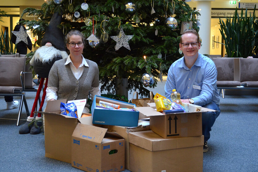 Nadine Abmeier und Marc Pingel vor dem Weihnachtsbaum des Krankenhauses mit den Paketen für die Johanniter-Weihnachtstrucker. 