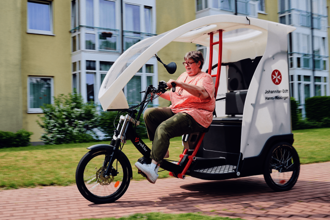 Eine Frau fährt ein E-Bike mit überdachtem Anhänger vor dem Johanniter-Stift Hannover-Ricklingen