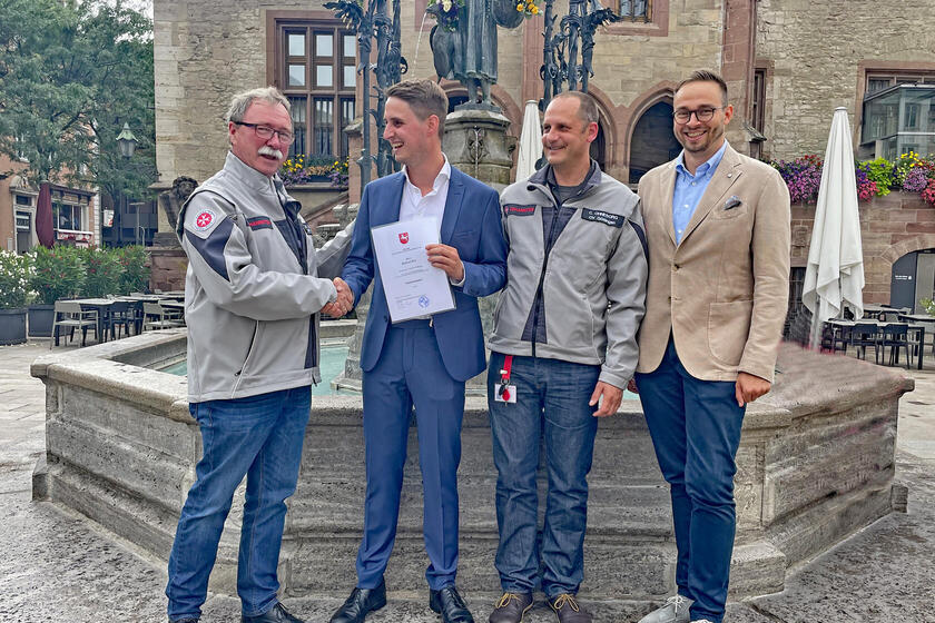 Vier Männer, zwei davon in Johanniter-Kleidung, vor dem Gänselieselbrunnen in Göttingen. Einer häte eine Urkunde hoch und ihm wird gratuliert.