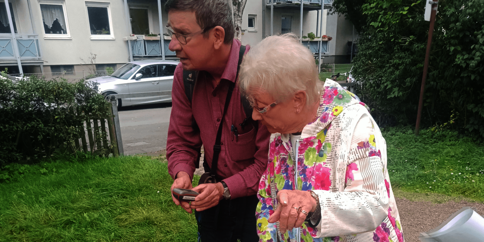 Zwei Personen im Freien. Ein Mann hält eine kleines Gerät in der Hand, eine ältere Frau neben ihm sieht darauf und deutet mit der Hand eine Richtung.