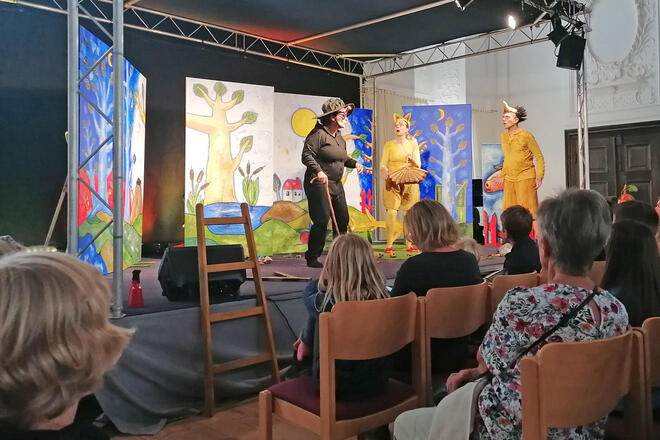 Menschen vor einer Theaterbühne. Auf der Bühne drei kostümierte Schauspieler.