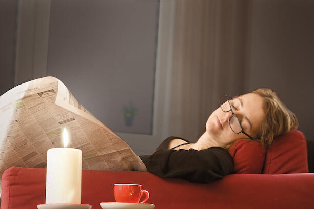 Frau liegt schlafend auf dem Sofa und einer Zeitung, die über eine brennende Kerze ragt, in der Hand.