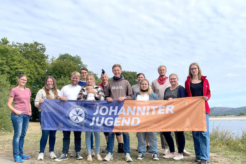Mehrere Teilnehmende stehen in zwei Reihen an der Küste eines Sees, zu ihrer linken. Die vorderen halten ein Johanniter-Jugend-Banner vor sich.
