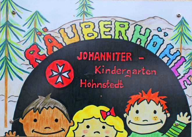 Türschild der Kita Räuberhöhle in Hohnstedt mit der Zeichungen von drei Kindern, dem Johanniter-Logo und einem kleinen Wald im Hintergrund. 